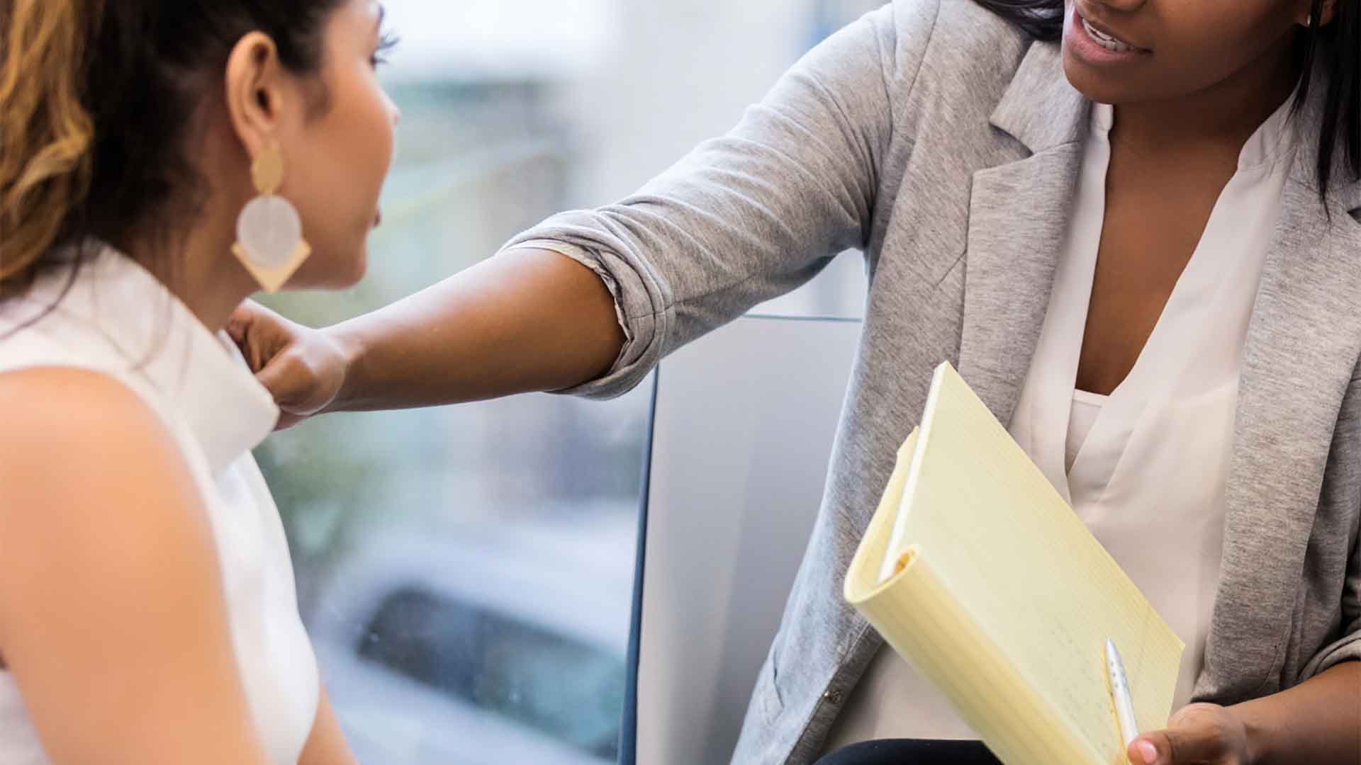 Primo piano di una counselor che posa una mano confortante sulla spalla di un'altra donna. La counselor, con in mano un blocco note giallo, parla con un'espressione empatica durante quella che sembra essere una seduta di terapia o di supporto in caso di crisi.