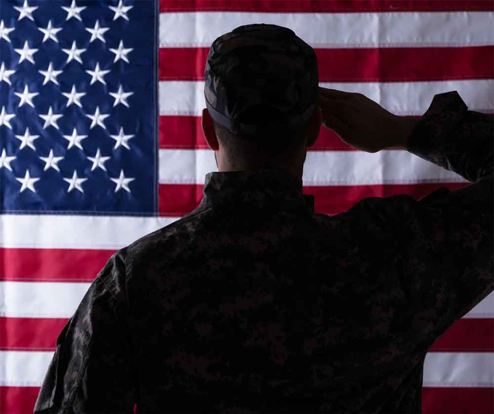 A soldier in uniform stands in silhouette, seen from behind, while saluting a large American flag.