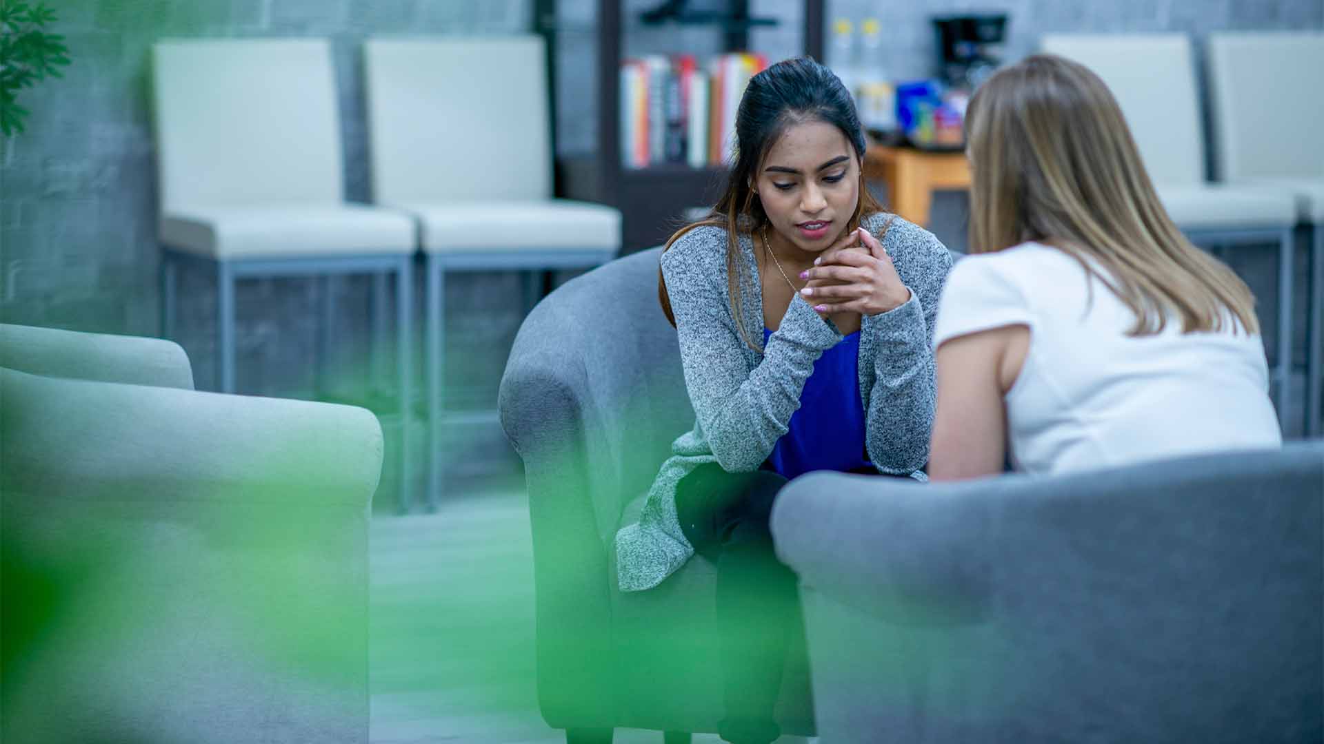 تتحدث شابة، بوجهٍ قلق، جالسةً على كرسيٍّ بذراعين خلال جلسة استشارية. يجلس مُعالِجٌ نفسيّ، يُرى من الخلف، في الجهة المقابلة ويستمع باهتمام.