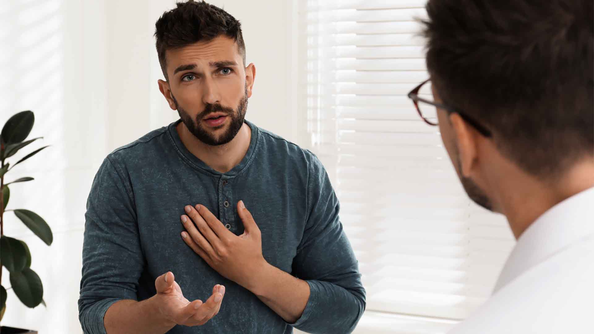 Un uomo con la barba parla con un'espressione seria durante una seduta di consulenza. Gesticola con le mani, una vicino al petto, mentre parla con un terapeuta, che è visto da dietro le spalle.