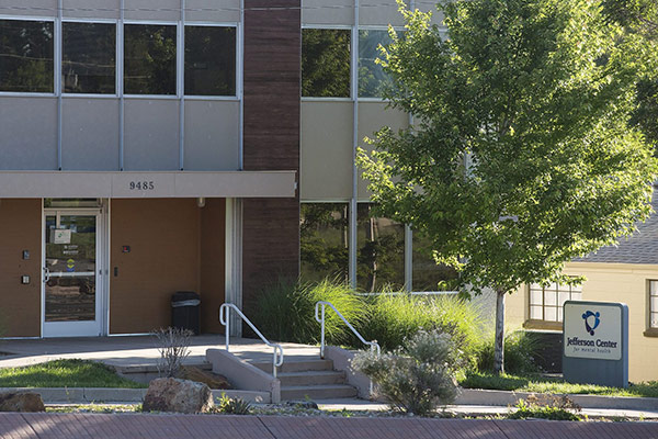 The exterior entrance of a multi-story office building at 9485 West Colfax Ave on a sunny day. A sign on the lawn to the right reads "Jefferson Center for Mental Health." 