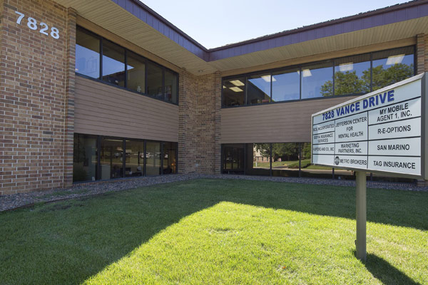 The exterior of a two-story, brick and brown-siding office building at 7828 Vance Drive on a sunny day. In the foreground, a white directory sign on a green lawn lists the tenants, including: Jefferson Center for Mental Health