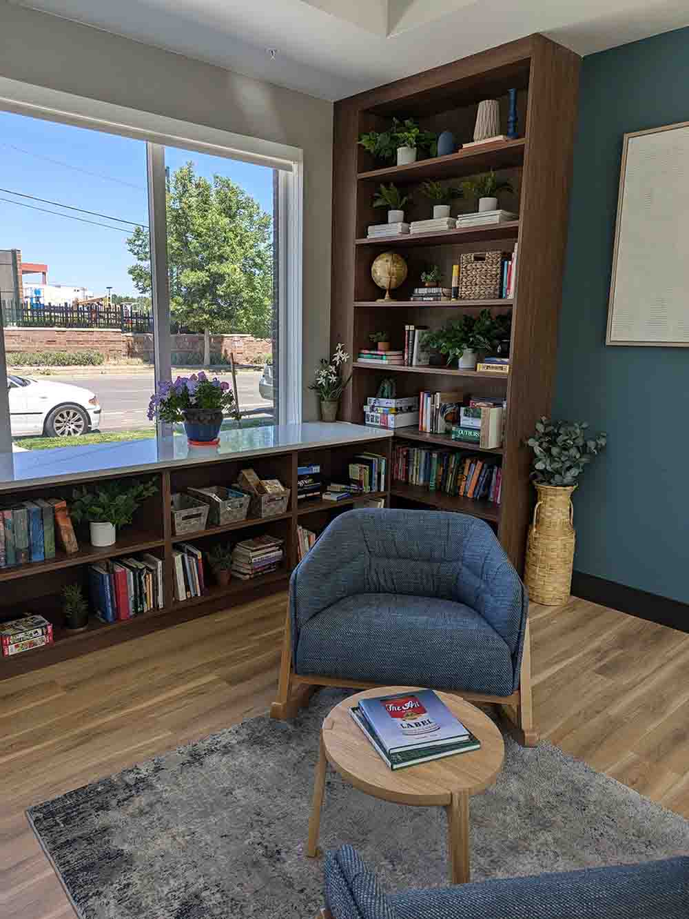 A cozy library and reading nook at the Solid Ground apartments. The space is furnished with a comfortable blue armchair on a patterned rug, surrounded by dark wood, built-in bookshelves filled with books and decorative plants. A large window lets in bright, natural light.