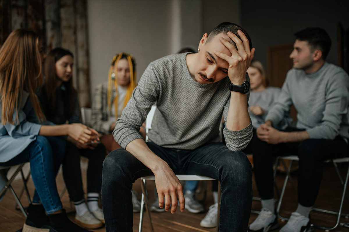 Um jovem está sentado em uma reunião de grupo de apoio, olhando para baixo com uma expressão angustiada e a mão na testa. Outros membros do grupo estão sentados em círculo ao seu redor, desfocados no fundo, concentrando a atenção em seu momento de vulnerabilidade.