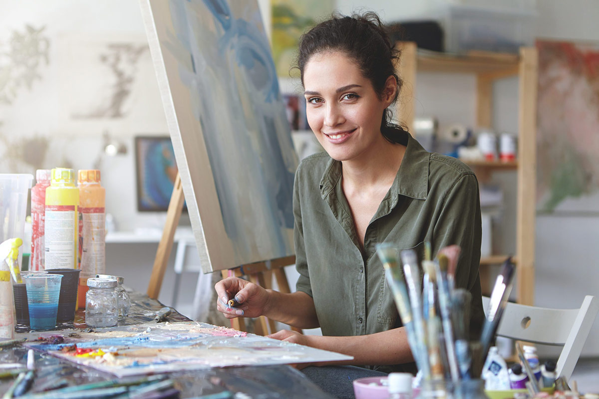A female artist smiles warmly at the camera while sitting in her bright art studio. She holds an artist's palette covered in colorful paint and a palette knife, and her work table is filled with brushes. A large, abstract painting on an easel is visible behind her.