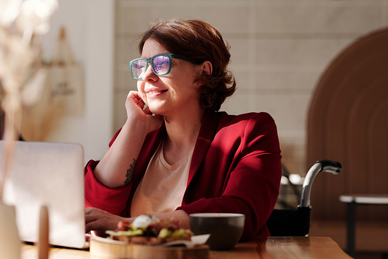 A smiling woman with short, reddish-brown hair and stylish blue-green glasses sits at a table in a sunlit cafe. She is wearing a red blazer and rests her chin on her hand while looking contentedly to the side