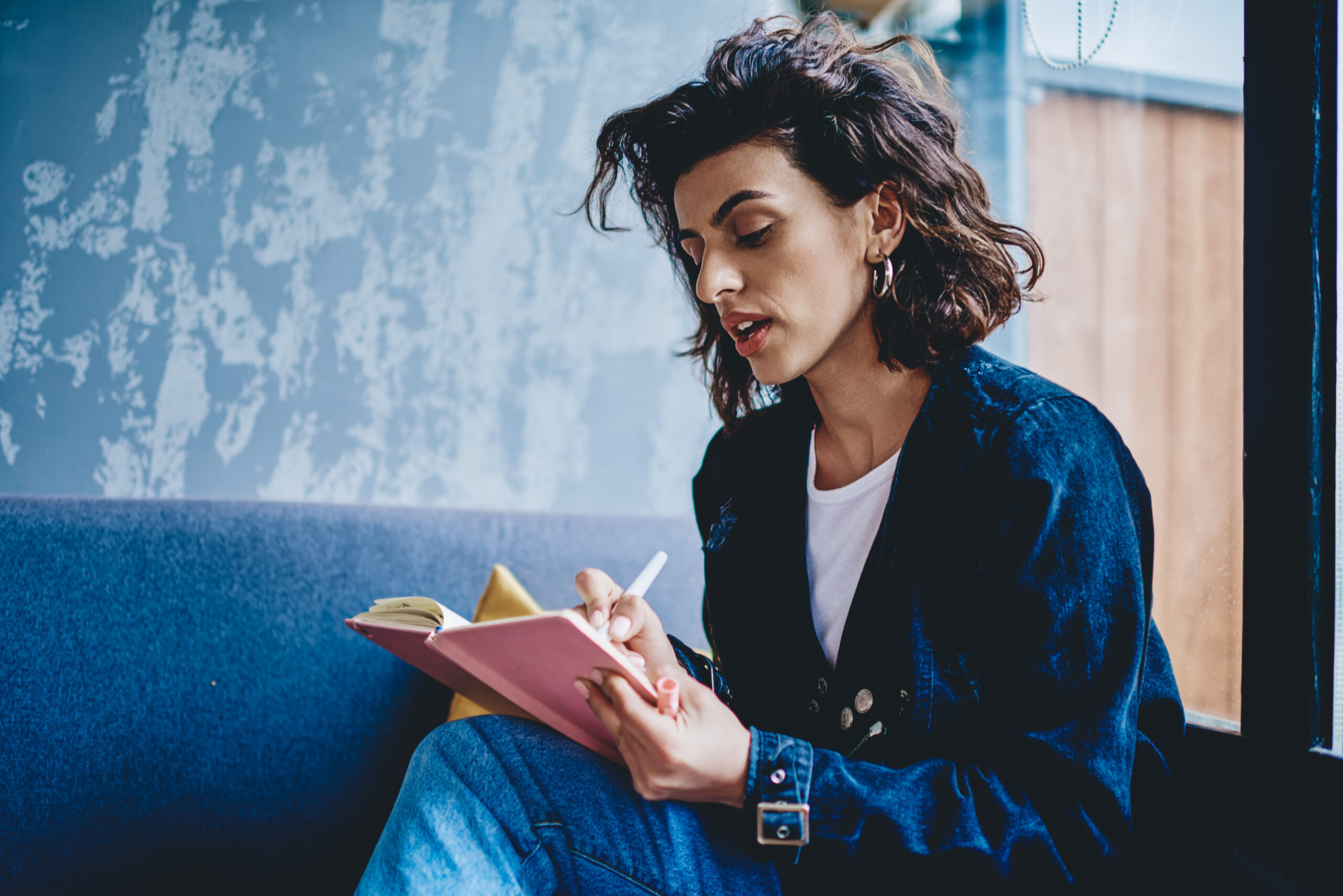 A young woman with wavy, dark hair sits by a window and writes in a pink notebook. She is wearing a denim jacket and has a focused, contemplative expression.