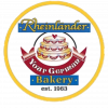Logo for Rheinländer Bakery. The circular logo features an illustration of a three-tiered decorated cake in the center. The name "Rheinländer Bakery" is written in gold on blue banners above and below the cake.
