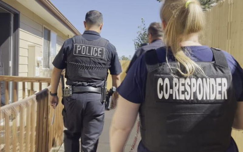 Seen from behind, a police officer and a female co-responder walk side-by-side along an outdoor walkway next to a building.
